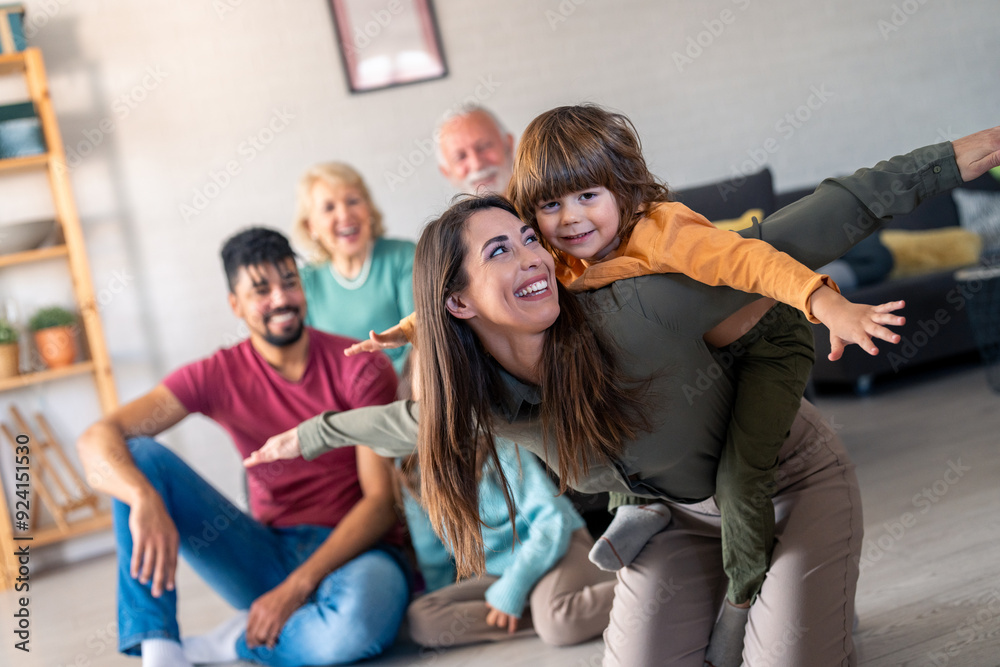 Obraz premium A joyful family with diverse ages smiles during a home photo session, featuring a mother playfully giving her son a piggyback ride while others sit on the sofa and floor.