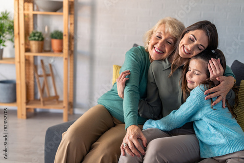 A joyful grandmother, mother, and young daughter sit closely on a couch, sharing a warm, affectionate embrace in a cozy living room.