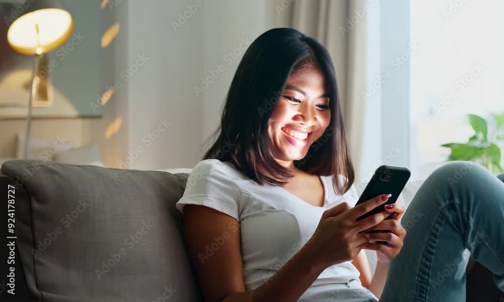 Multicultural young adult female sitting and laughing on sofa, typing on cellphone. Modern apartment, leisure living in daytime Multicultural young adult female sitting and laughing on sofa, typing on cellphone. Modern apartment, leisure living in daytime