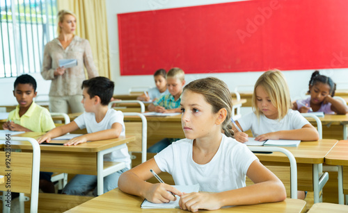 Diligent elementary school student tween girl studying with classmates, making notes of teacher lecture