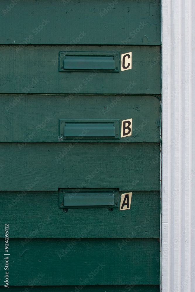 The exterior wall of a green wooden apartment building has three mail slots. There are letters, a,b, and c, next to each slot. The trim on the edge of the wall is white. The postal slots are retro. The exterior wall of a green wooden apartment building has three mail slots. There are letters, a,b, and c, next to each slot. The trim on the edge of the wall is white. The postal slots are retro.