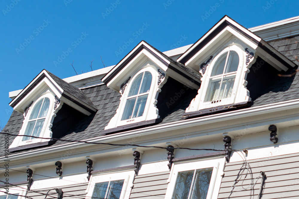 Three peaked dormer-style windows on a hip roof covered with black shingles. The vintage white wooden frames have arch-shaped glass and closed single-hung panes with white curtains hanging inside.  