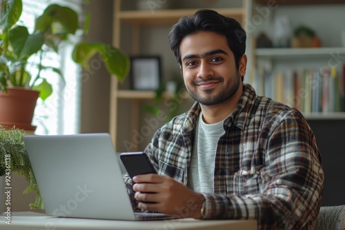 Young adult Arabic businessman holding cellphone smartphone at hands, sitting at desk with laptop computer at home office. Smiling Indian man using phone application, Generative AI