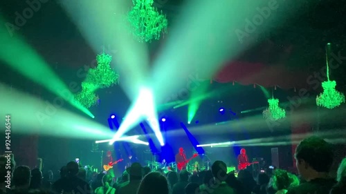 An indoor concert with dramatic stage lighting. A rock band performs on a stage at an indoor concert venue, with dramatic lighting on stage and spreading over the crowd.
