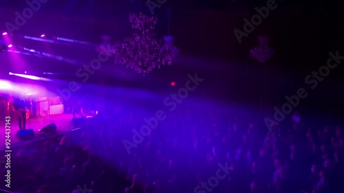 An indoor concert with dramatic stage lighting. A rock band performs on a stage at an indoor concert venue, with dramatic lighting on stage and spreading over the crowd.
