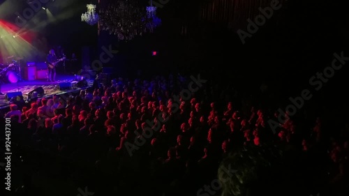 An indoor concert with dramatic stage lighting. A rock band performs on a stage at an indoor concert venue, with dramatic lighting on stage and spreading over the crowd.