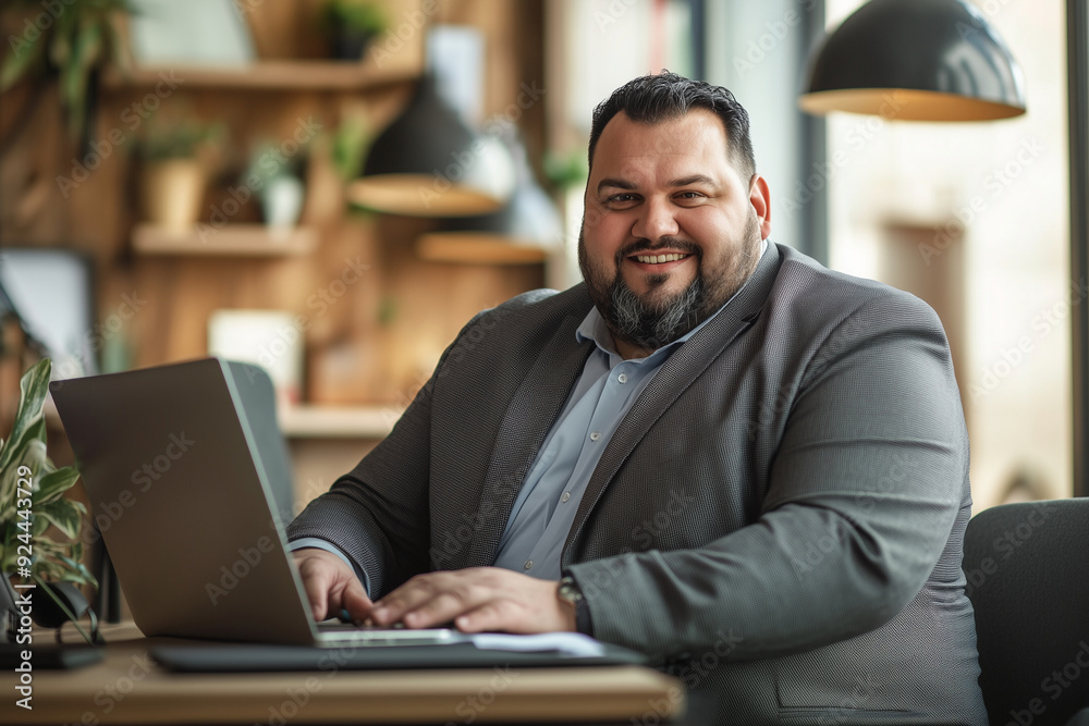 Plus size man in suit is sitting at a desk with a laptop. He is smiling and he is happy. Plus size man in suit is sitting at a desk with a laptop. He is smiling and he is happy.