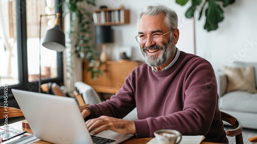 An elderly man smiling and holding glasses while sitting at an open laptop on the table of a home office