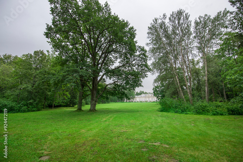 A vibrant green meadow stretches out under a cloudy sky, framed by tall trees and leading to a distant historic estate, evoking tranquility and natural beauty.