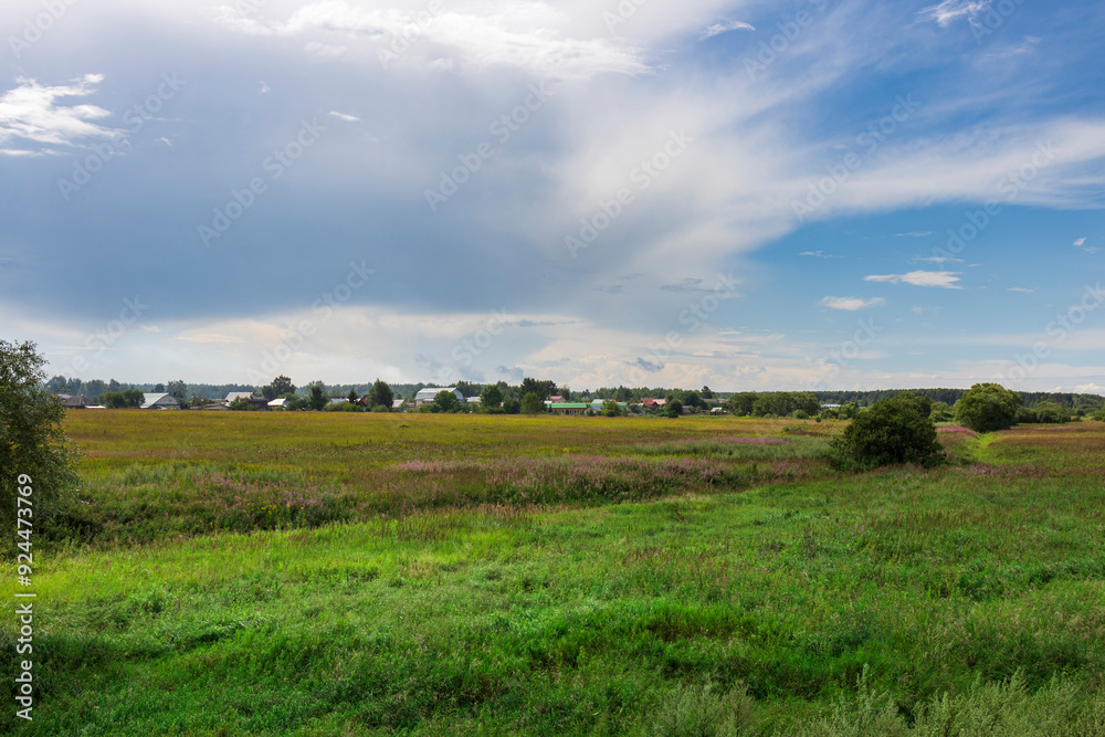 Lush green fields stretch towards a small town, illuminated by the golden hues of the late afternoon sun against a backdrop of soft, fluffy clouds. Lush green fields stretch towards a small town, illuminated by the golden hues of the late afternoon sun against a backdrop of soft, fluffy clouds.
