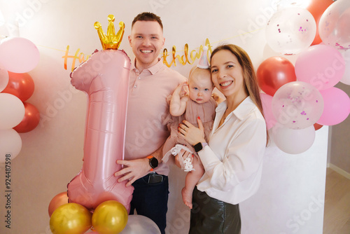 Happy and gentle parents hold baby daughter with birthday balloon decor.