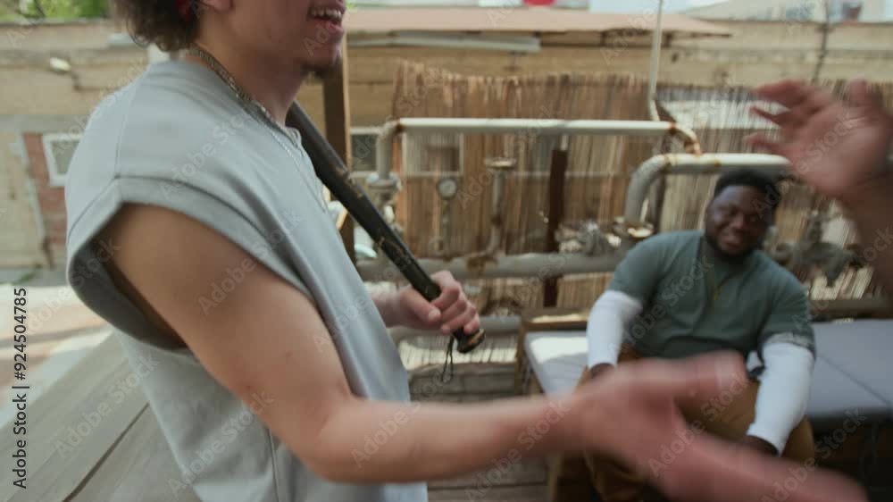 Young man holding baseball bat and talking with buddy, then greeting Black friend joining them on urban rooftop Young man holding baseball bat and talking with buddy, then greeting Black friend joining them on urban rooftop