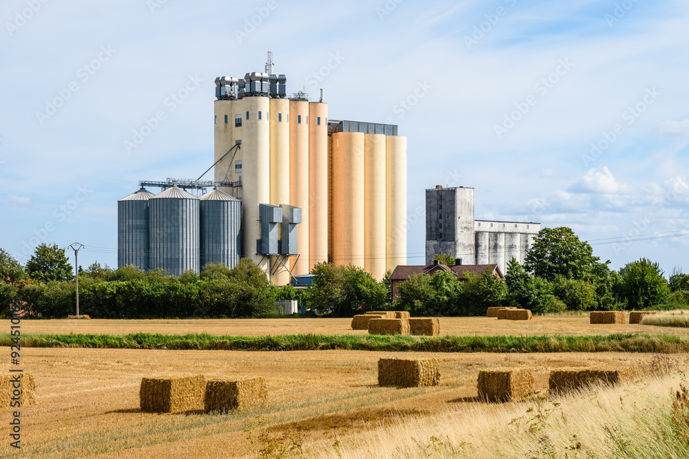 Two grain tower silos in the countryside, a smaller and older gray one next to a more recent and much larger coloured one, with bales of straw in the foreground on a sunny summer day. Two grain tower silos in the countryside, a smaller and older gray one next to a more recent and much larger coloured one, with bales of straw in the foreground on a sunny summer day.