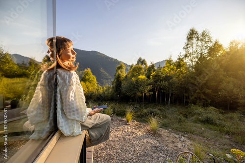 A woman sits quietly by a window, gazing at the surrounding mountains and forest. The serene landscape and reflection create a perfect moment of solitude and connection with nature