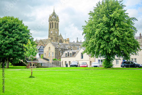 Public park along the banks of the river Tweed in Peeples a village in the Borders region of Scotland with parked cars and a stone church spire in the background. 