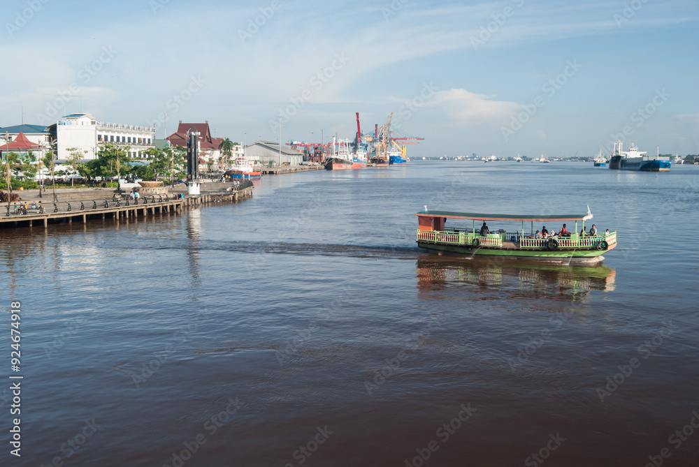 Fototapeta premium A traditional riverboat glides along a bustling urban harbor, showcasing a blend of modern industry and local culture with waterfront buildings and docks.