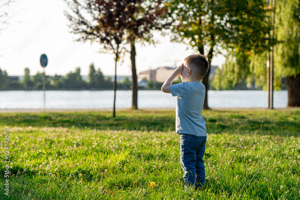 A little toddler boy of three and a half years old is standing in a summer park near a lake with his eyes closed. Childhood and nature. A little toddler boy of three and a half years old is standing in a summer park near a lake with his eyes closed. Childhood and nature.
