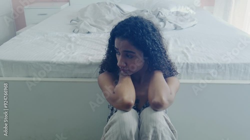 A wide shot of the young curly-haired pensive lonely anxious Indian Asian female or woman sitting on the floor in destress in the bedroom. Concept of heartbreak, depression, mental health medication