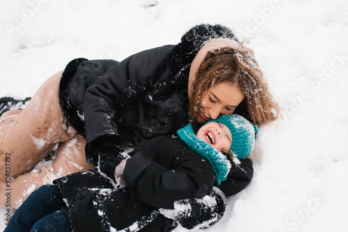 Happy woman and child are playing in the snow. Mom is laying on the ground and son is laying on top of her. They are both smiling and enjoying the moment photo