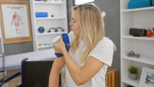 A blonde woman exercises her arm with a dumbbell in a clinic's rehab room, surrounded by medical equipment.