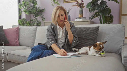 A mature woman with blonde hair sits in a cozy living room, talking on the phone while writing in a notebook, with a small dog playing on the couch beside her.