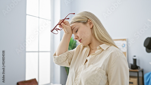 A thoughtful young woman with blonde hair holding glasses in a bright modern office.