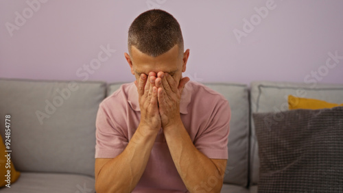 Fotografie Young man sitting on a sofa at home covering his face with both hands, in a moment of apparent stress