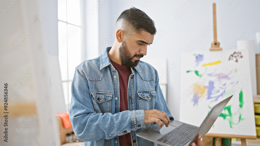 A young bearded man in a denim jacket uses a laptop in a brightly lit art studio with paintings in the background. A young bearded man in a denim jacket uses a laptop in a brightly lit art studio with paintings in the background.