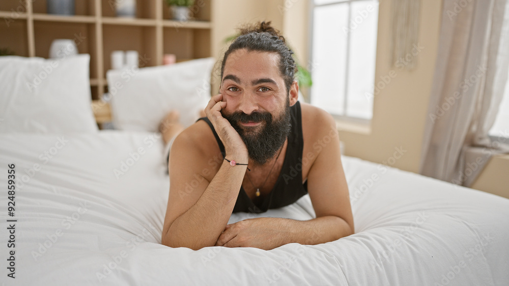 Handsome bearded hispanic man lying on a bed in a bright bedroom, smiling casually in a relaxed pose Handsome bearded hispanic man lying on a bed in a bright bedroom, smiling casually in a relaxed pose