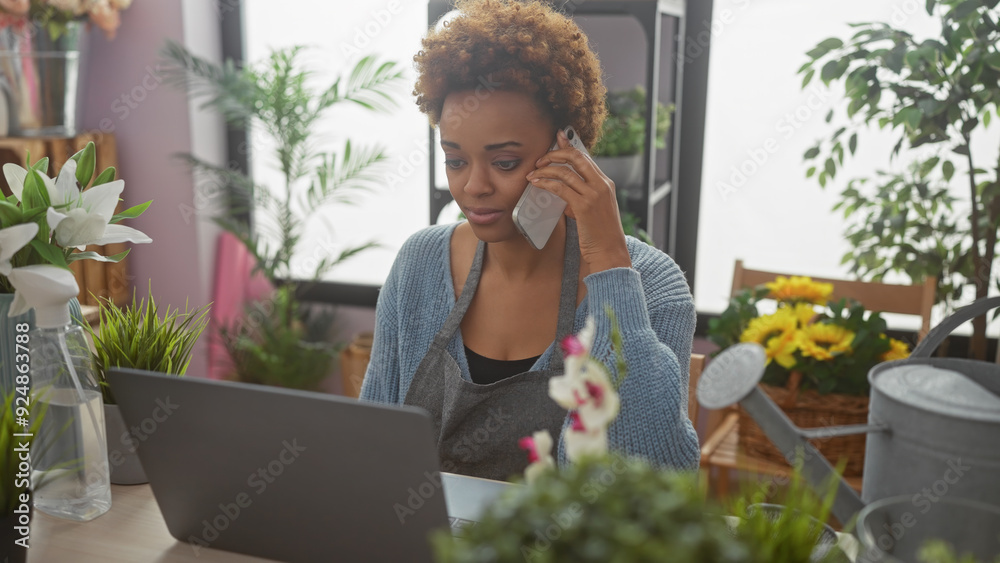 African american woman talking on the phone in a flower shop with plants and laptop, depicting entrepreneurship and multitasking. African american woman talking on the phone in a flower shop with plants and laptop, depicting entrepreneurship and multitasking.