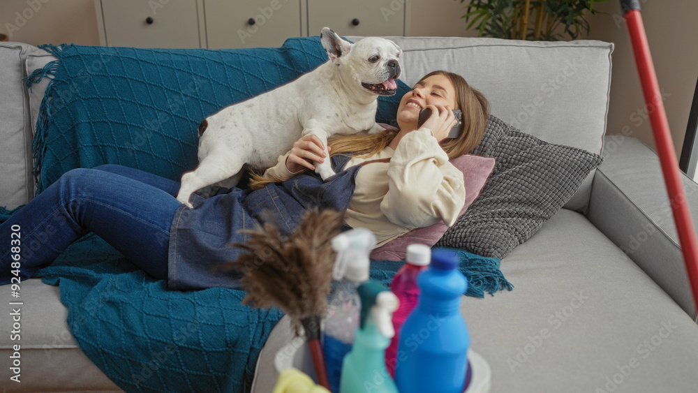 A young beautiful blonde woman talking on the phone while lying on her living room couch with her pet canine in an apartment, surrounded by cleaning supplies. A young beautiful blonde woman talking on the phone while lying on her living room couch with her pet canine in an apartment, surrounded by cleaning supplies.
