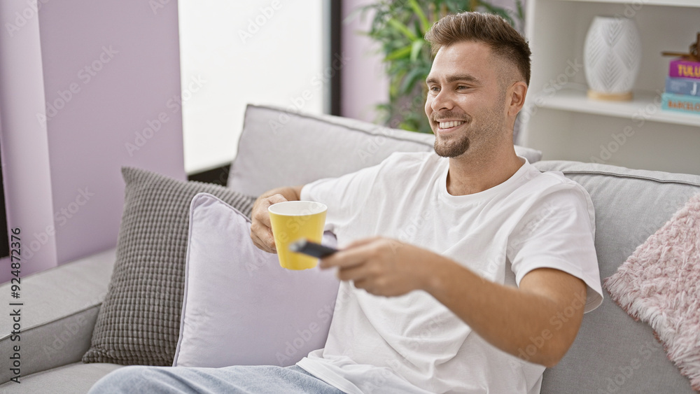 A relaxed young hispanic man with a beard enjoying coffee while watching tv in a modern living room. A relaxed young hispanic man with a beard enjoying coffee while watching tv in a modern living room.