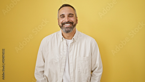 A smiling hispanic senior man with a beard and grey hair stands against a yellow isolated background wearing a white shirt.