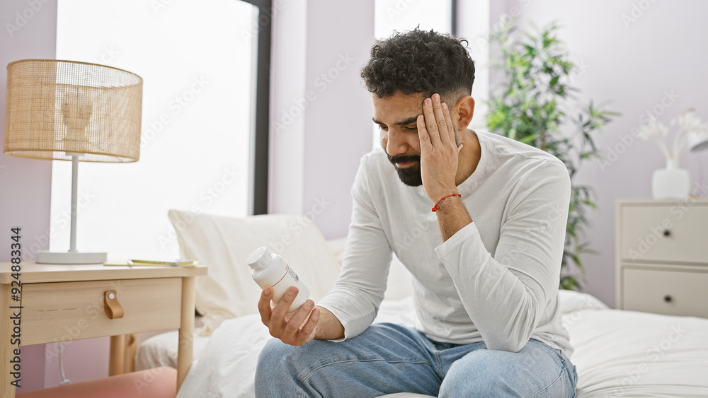 A young hispanic man with a beard looks concerned at a pill bottle while sitting on a bed indoors A young hispanic man with a beard looks concerned at a pill bottle while sitting on a bed indoors