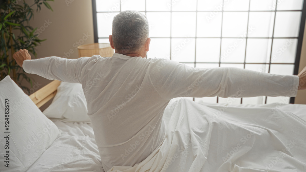 Fototapeta premium Middle-aged man stretching on bed in bedroom of home with bright lighting and modern interior design.
