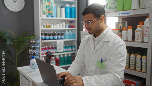 Wallpaper Mural Young hispanic man using a laptop in a pharmacy with shelves full of various products and medicines around. Torontodigital.ca