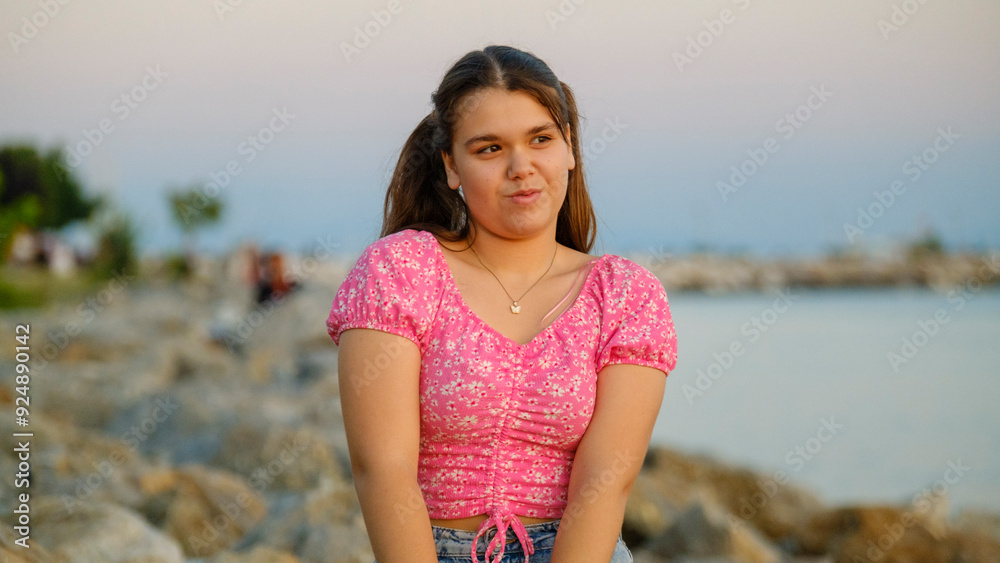 A chubby teenage girl sitting on the rocks by the seaside on a sunny summer day, smiling and looking at the camera