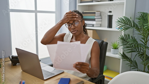 An adult african american woman with curly hair looks stressed while reading documents in a modern office.