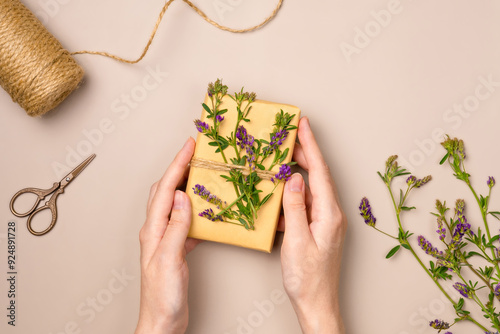Female's hands holding gift box with flowers on beige background from above. Gift wrapping composition. Flat lay, top view.