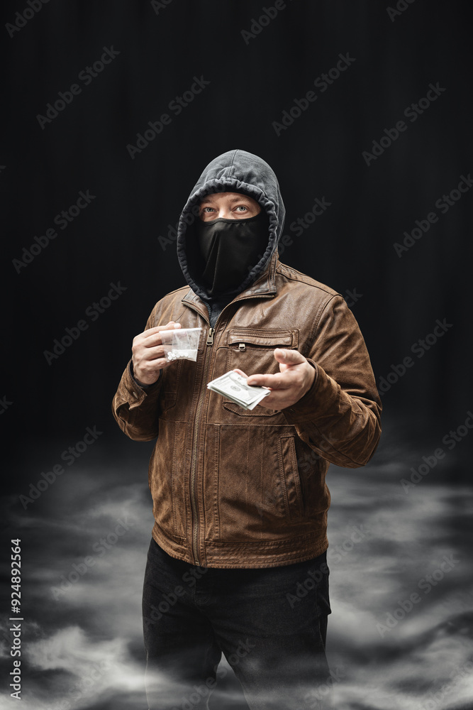 Drug dealer, dangerous hooded criminal man in black mask and brown leather jacket holding transparent plastic bag with drugs and dollar bills, on a dark background with smoke Drug dealer, dangerous hooded criminal man in black mask and brown leather jacket holding transparent plastic bag with drugs and dollar bills, on a dark background with smoke