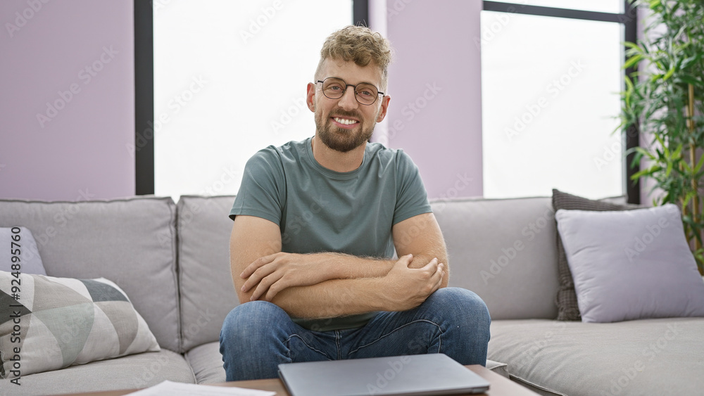 A smiling bearded caucasian man with blue eyes sits casually at home on a gray sofa, portraying a relaxed and welcoming atmosphere. A smiling bearded caucasian man with blue eyes sits casually at home on a gray sofa, portraying a relaxed and welcoming atmosphere.