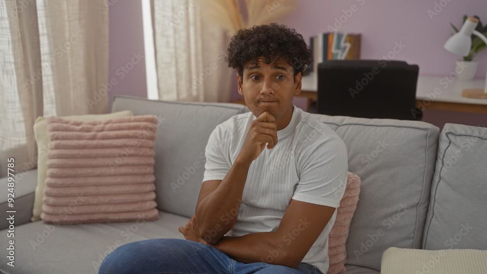 Young man sitting on a couch in a cozy living room, contemplating deeply with a thoughtful expression in a modern apartment setting. Young man sitting on a couch in a cozy living room, contemplating deeply with a thoughtful expression in a modern apartment setting.