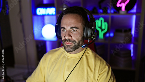 Portrait of a focused hispanic man with a beard wearing headphones in a colorful gaming room during night time. photo