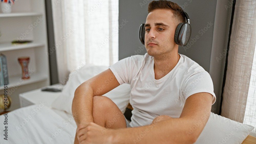 Portrait of a relaxed young man sitting on a bed with headphones in a bright bedroom at home. Portrait of a relaxed young man sitting on a bed with headphones in a bright bedroom at home.