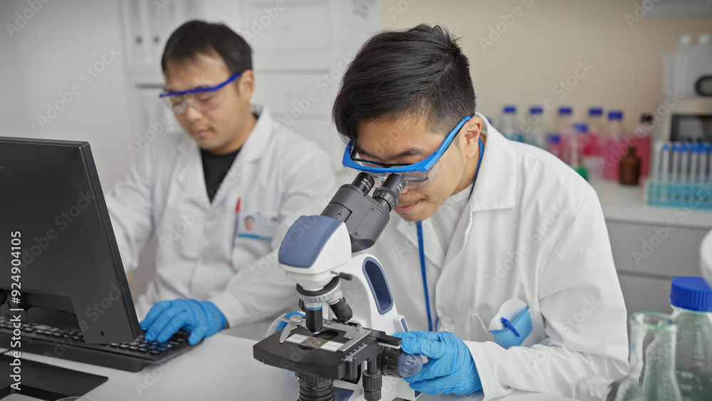 Two men working collaboratively in a modern laboratory, one using a microscope and another on a computer. Two men working collaboratively in a modern laboratory, one using a microscope and another on a computer.