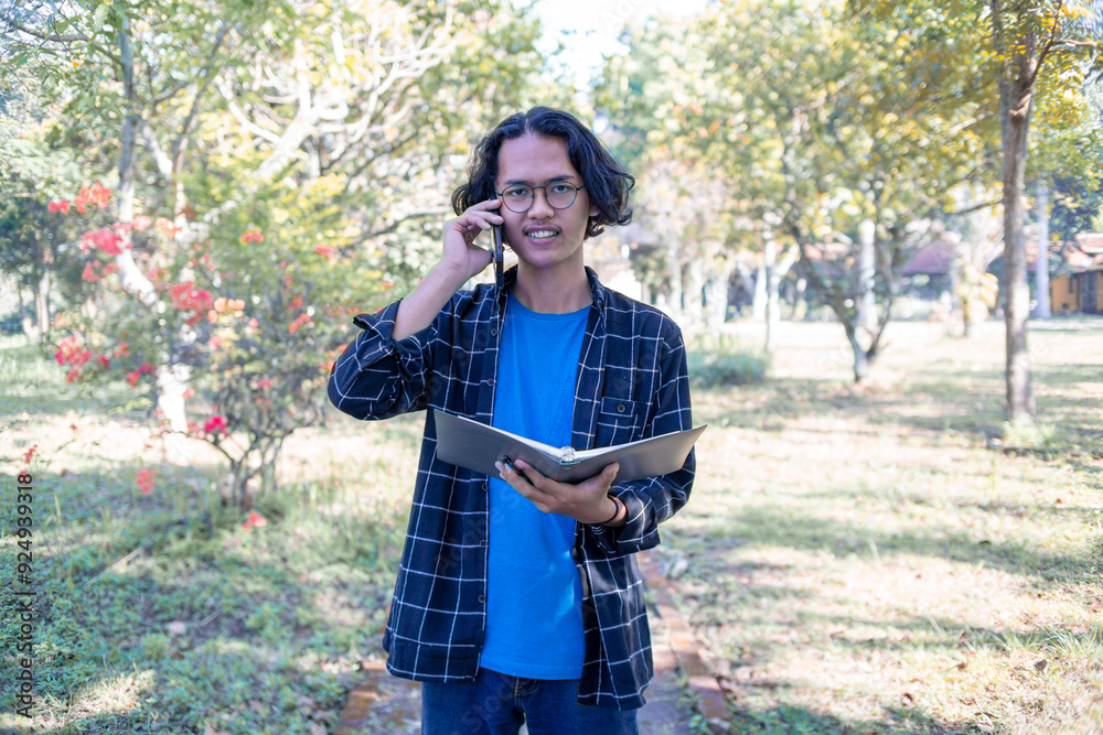 A male student wearing a plaid shirt and glasses is standing in the park answering a phone call and holding a book. A male student wearing a plaid shirt and glasses is standing in the park answering a phone call and holding a book.
