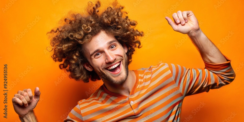 Amusing male model with curly hair and expressive facial gestures strikes a playful pose in front of a vibrant orange studio backdrop.