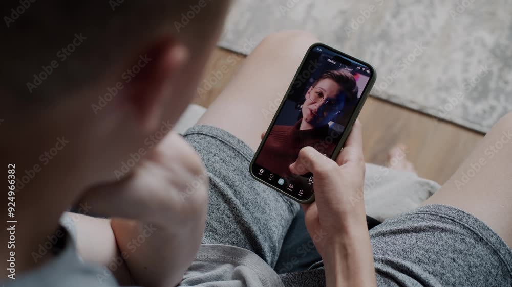Teen boy scrolls through and watches social network feed with short videos using mobile phone while sitting on sofa. Smartphone app with various video content displayed on phone screen. Close Up.