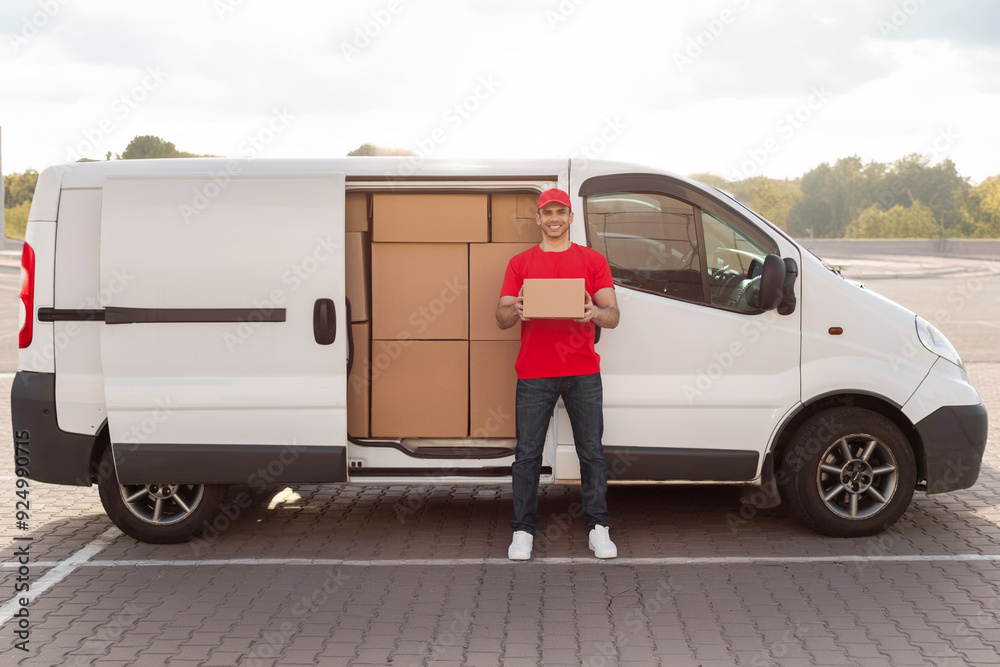 Young courier man in uniform holding deliver parcel and smiling at camera, standing near van, full length shot Young courier man in uniform holding deliver parcel and smiling at camera, standing near van, full length shot