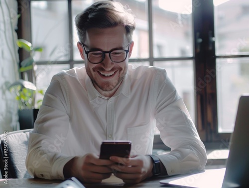 A smiling, glasses-wearing man in a shirt, seated at a desk using his cell phone. He seems pleased and connected in the digital age.